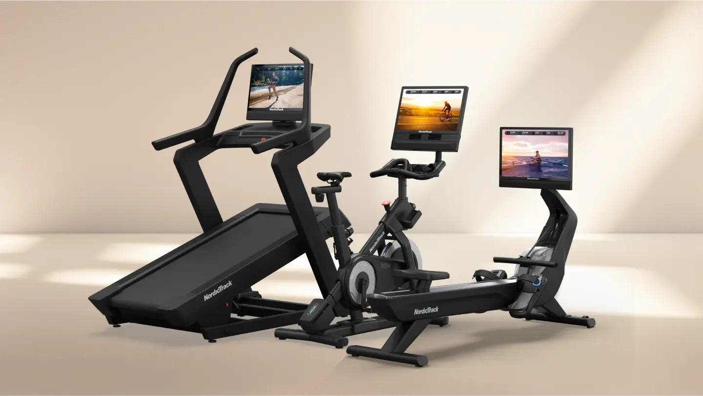 Black exercise equipment including a treadmill, exercise bike, and recumbent bike, all equipped with screens showing people exercising outdoors, placed against a beige wall on a light-colored floor.