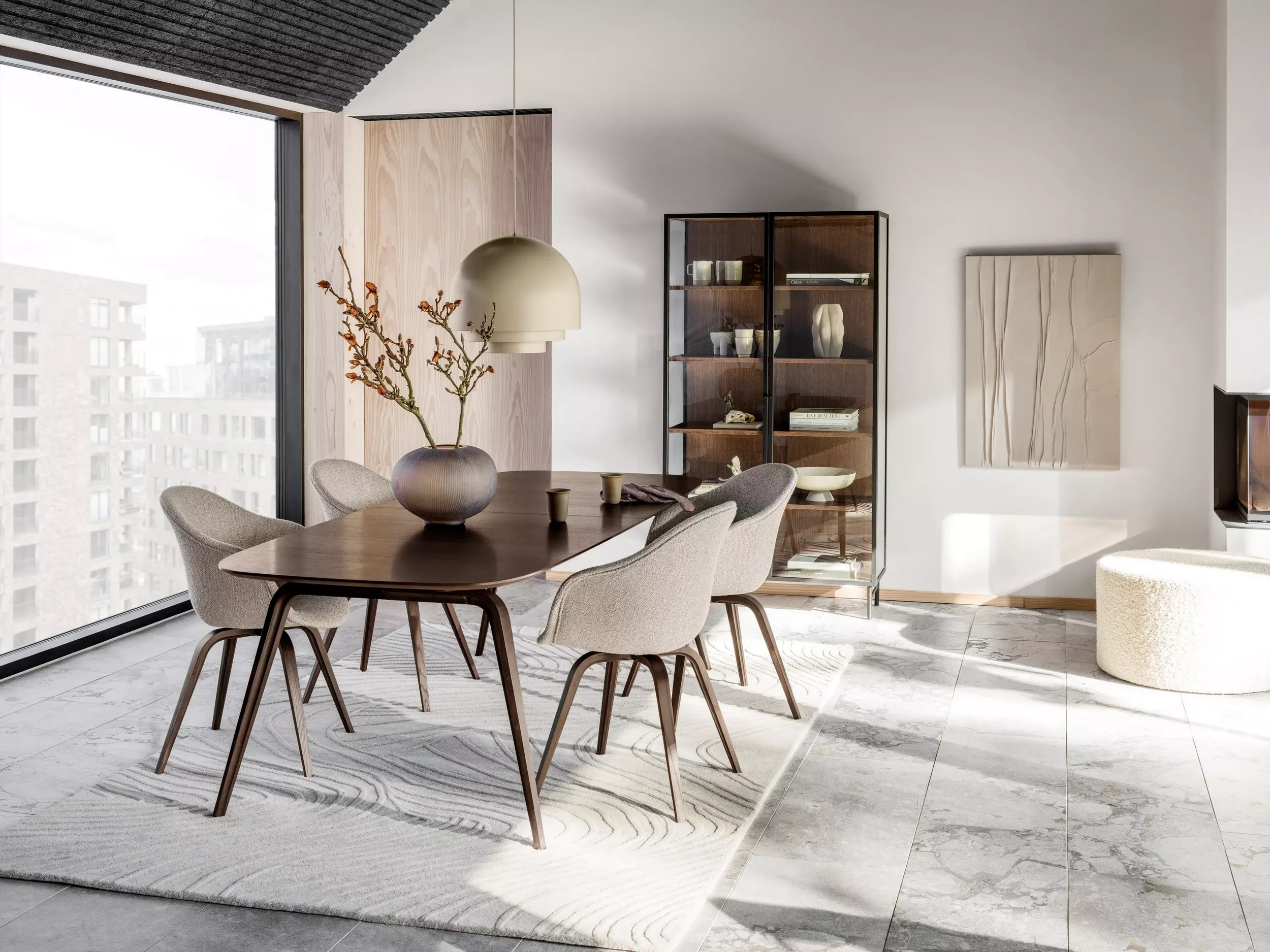 Modern dining room with a wooden table, six beige upholstered chairs, a large vase with branches on the table, a hanging light fixture, a glass display cabinet, textured artwork, and a white ottoman, with large windows and natural light.