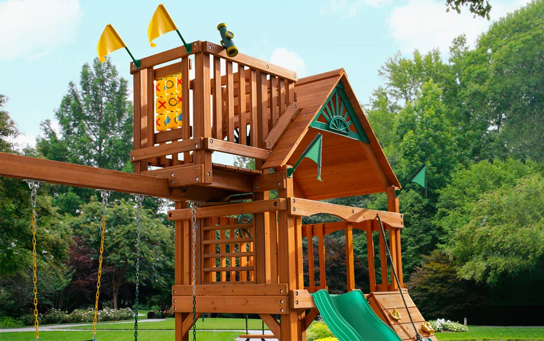 A wooden outdoor playset with slides, swings, a small house, and flags, set in a green park.