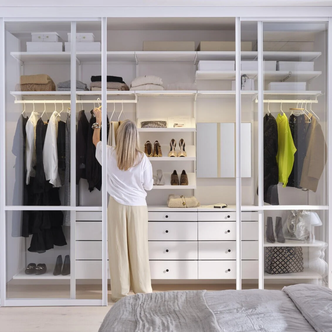 Woman organizing clothes in a white closet with shelves, drawers, and hanging space filled with clothing, shoes, and accessories.