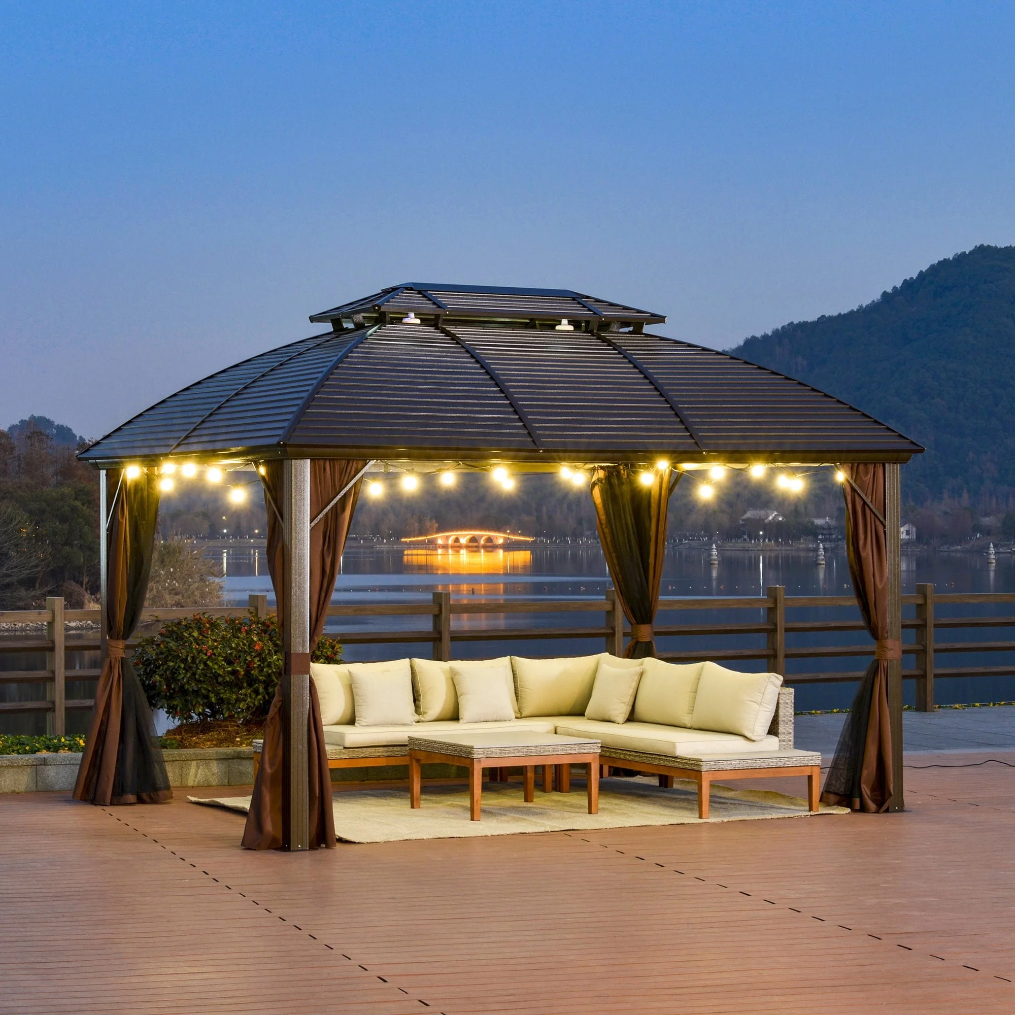 A gazebo with string lights, beige outdoor sectional sofa, and small coffee table on a wooden deck overlooking a river at dusk, with a lit bridge and mountains in the background.
