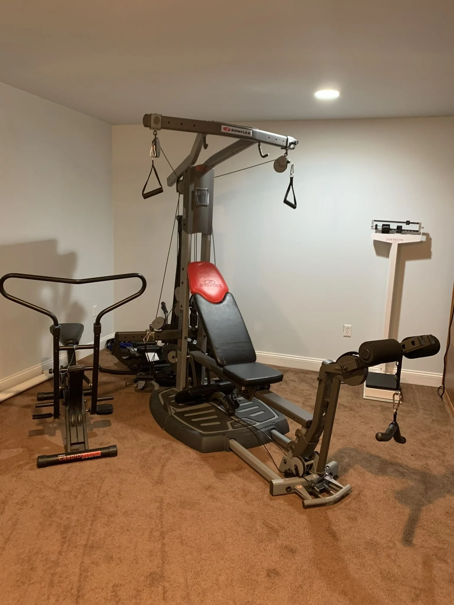 Home gym with a multi-purpose exercise machine, a recumbent exercise bike, and a scale against the wall.