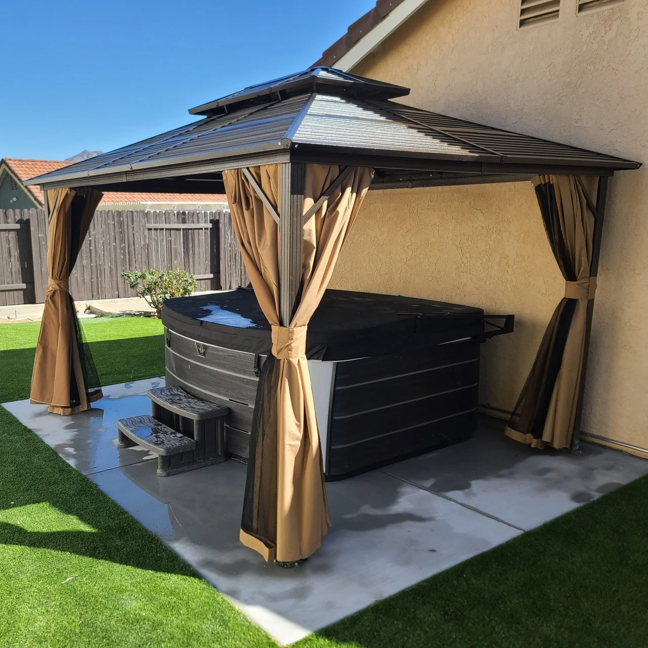 Outdoor hot tub under a metal gazebo with tan curtains, on a concrete slab, with a grassy yard and wooden fence in the background.