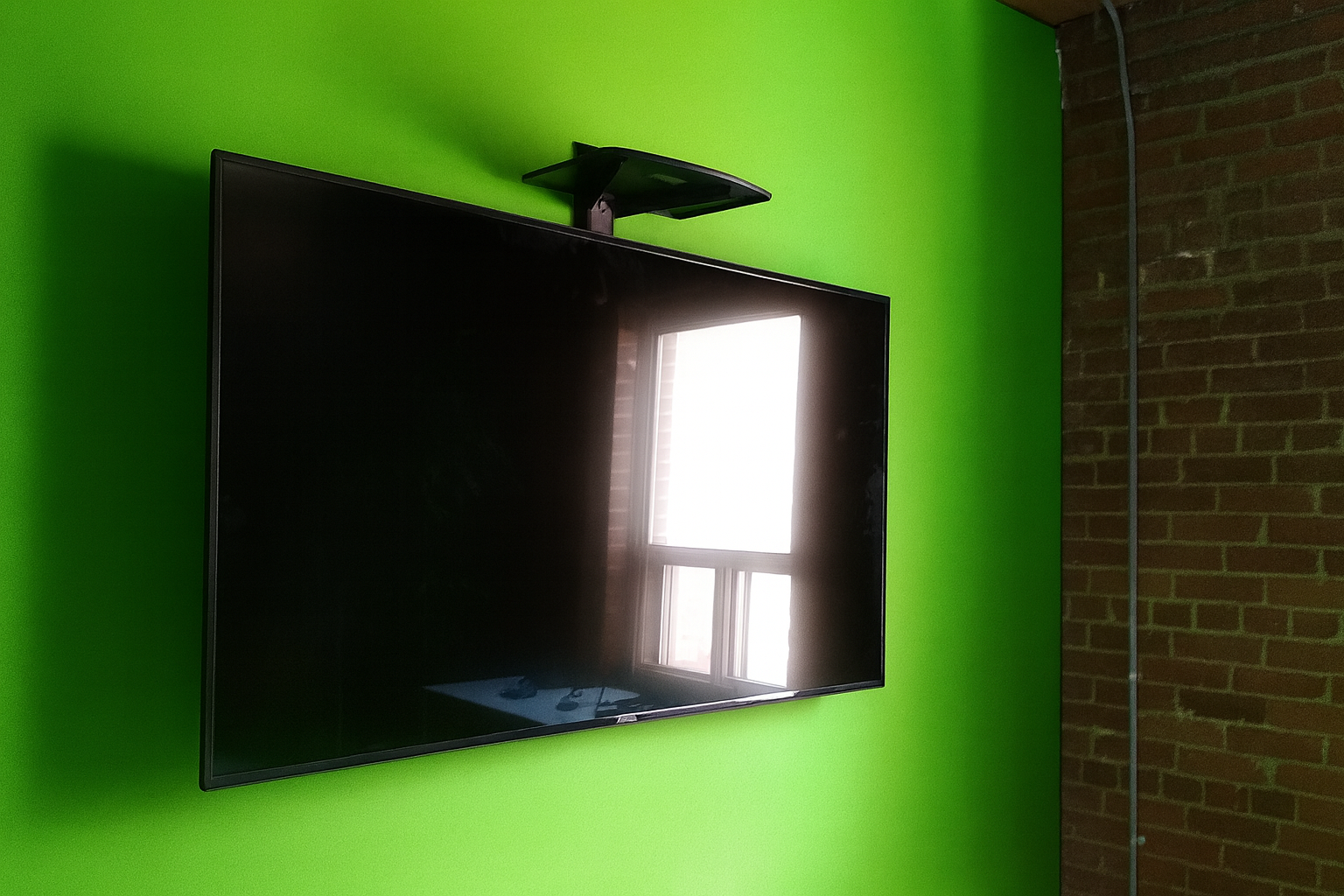 Flat-screen TV mounted on a bright green wall next to a window with a view of a brick wall outside.