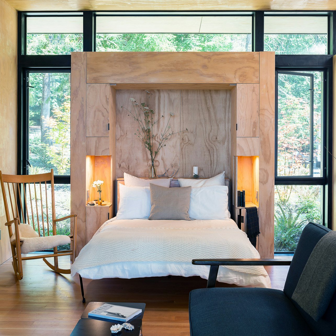 A bedroom with a large bed, wooden headboard, and large windows showing trees outside. The room has minimal decor with a cozy and modern style.