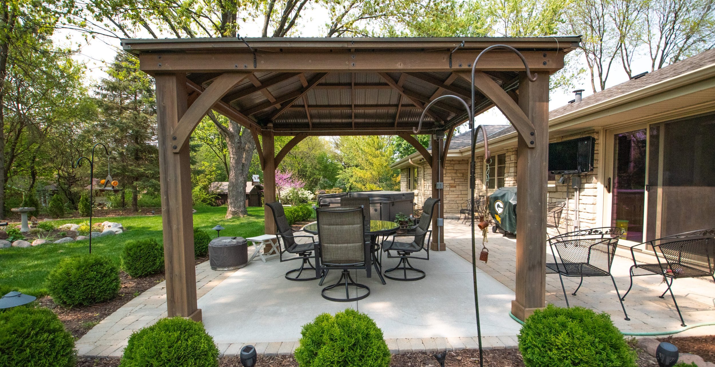 Backyard patio with a wooden pergola, metal chairs, a glass table, a hot tub, a TV, and surrounding garden with trees and bushes.