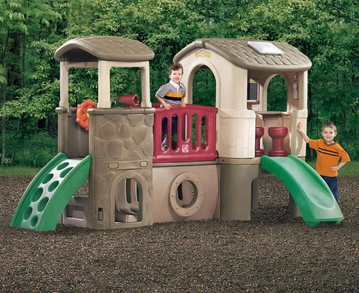 Children playing on a plastic playground structure resembling a treehouse, with slides, a steering wheel, and a small indoor area, set in a park with green trees in the background.