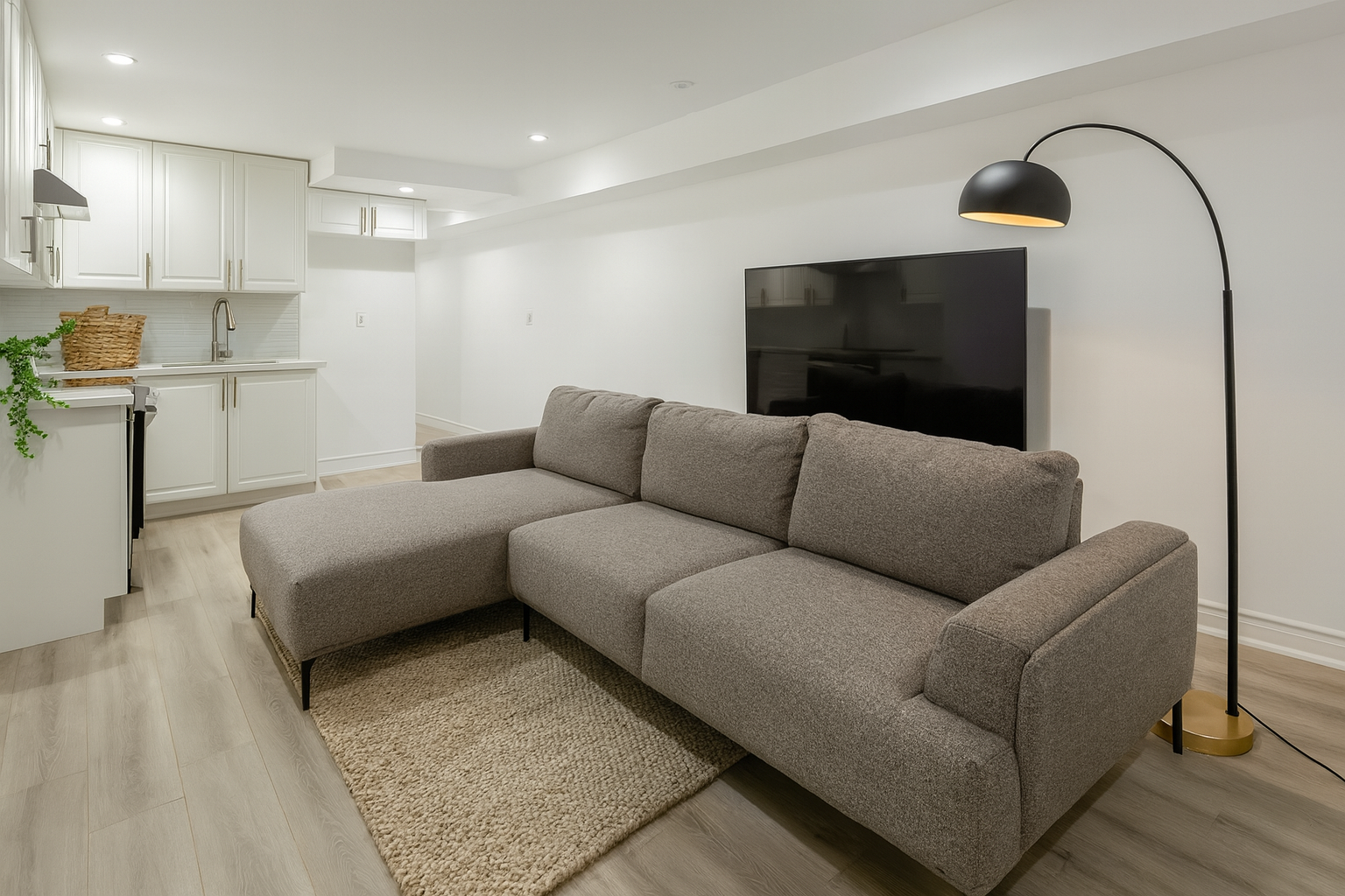 Modern living room with a gray sectional sofa, a flat-screen TV on the wall, and a black arc floor lamp. The background features a white kitchen with cabinetry and a small countertop.
