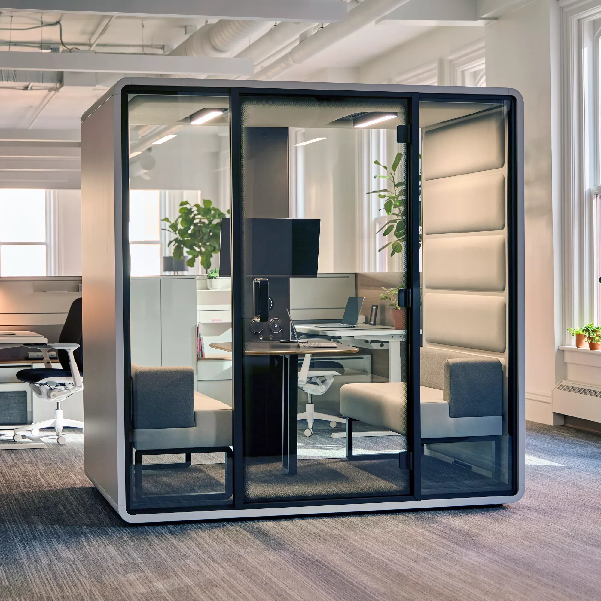 Modern office space with transparent glass private pod containing a desk, computer, and chairs, surrounded by other office furniture and potted plants.