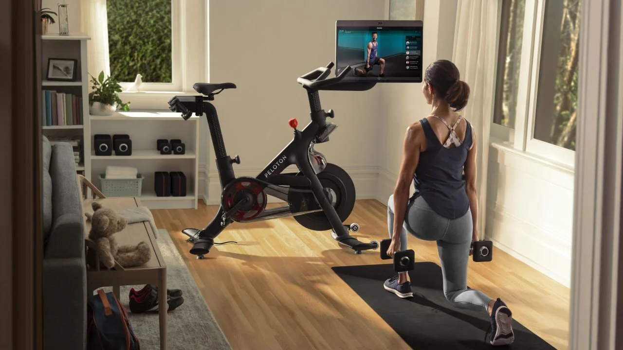 Woman working out at home with dumbbells on a yoga mat, watching a virtual trainer on a TV, with an exercise bike nearby and a shelf with books and decor in the background.