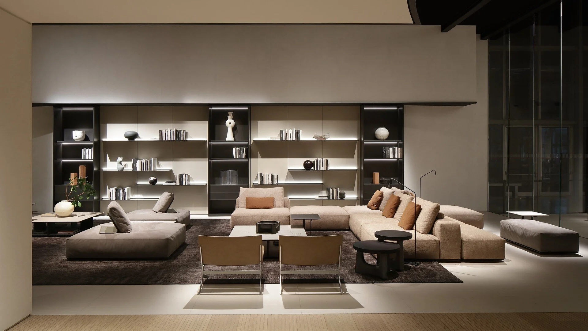 Modern living room with beige sectional sofa, armchair, and black coffee tables, backed by a built-in black and beige shelving unit with decorative objects and books, illuminated with ambient lighting.