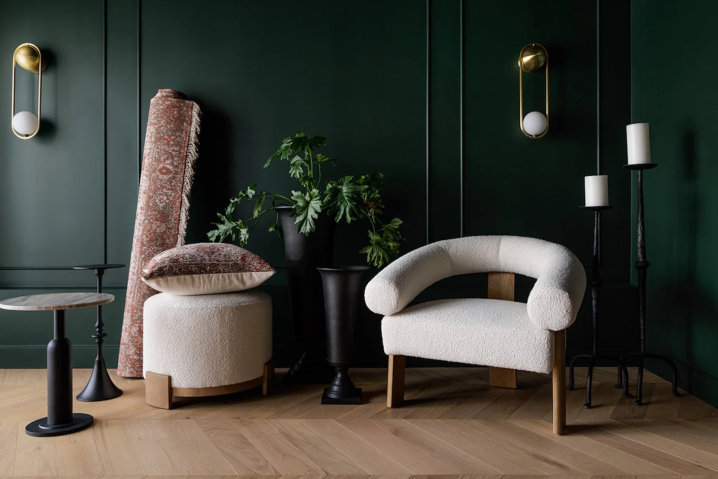 Living room corner with green wall, white textured armchair, beige ottoman with pink pillow, black side tables, potted green plant, and modern wall lights