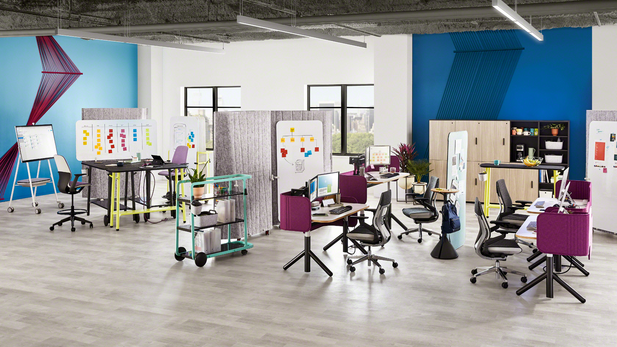 Open office space with desks, chairs, whiteboards, and dividers, featuring colorful accents and windows with city views.
