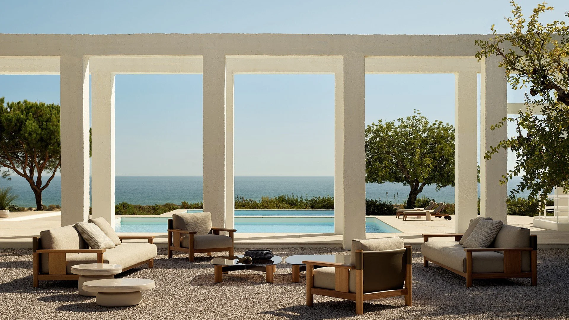 Outdoor living space with beige sofas and armchairs, a round coffee table, with a view of the ocean, trees, a pool, and lounge chairs in a modern, open structure