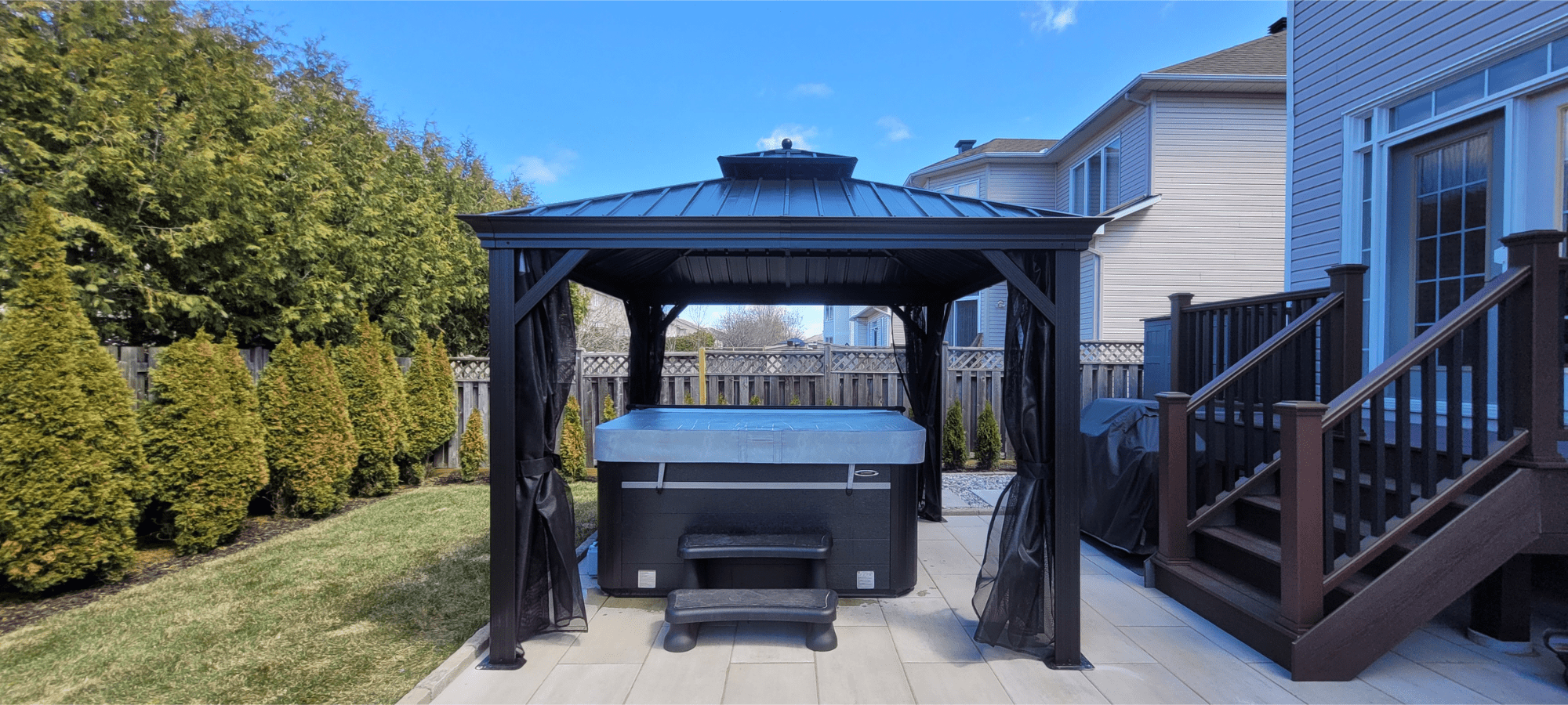 Backyard with a hot tub under a black gazebo, wooden stairs, and a fence with trees in the background.