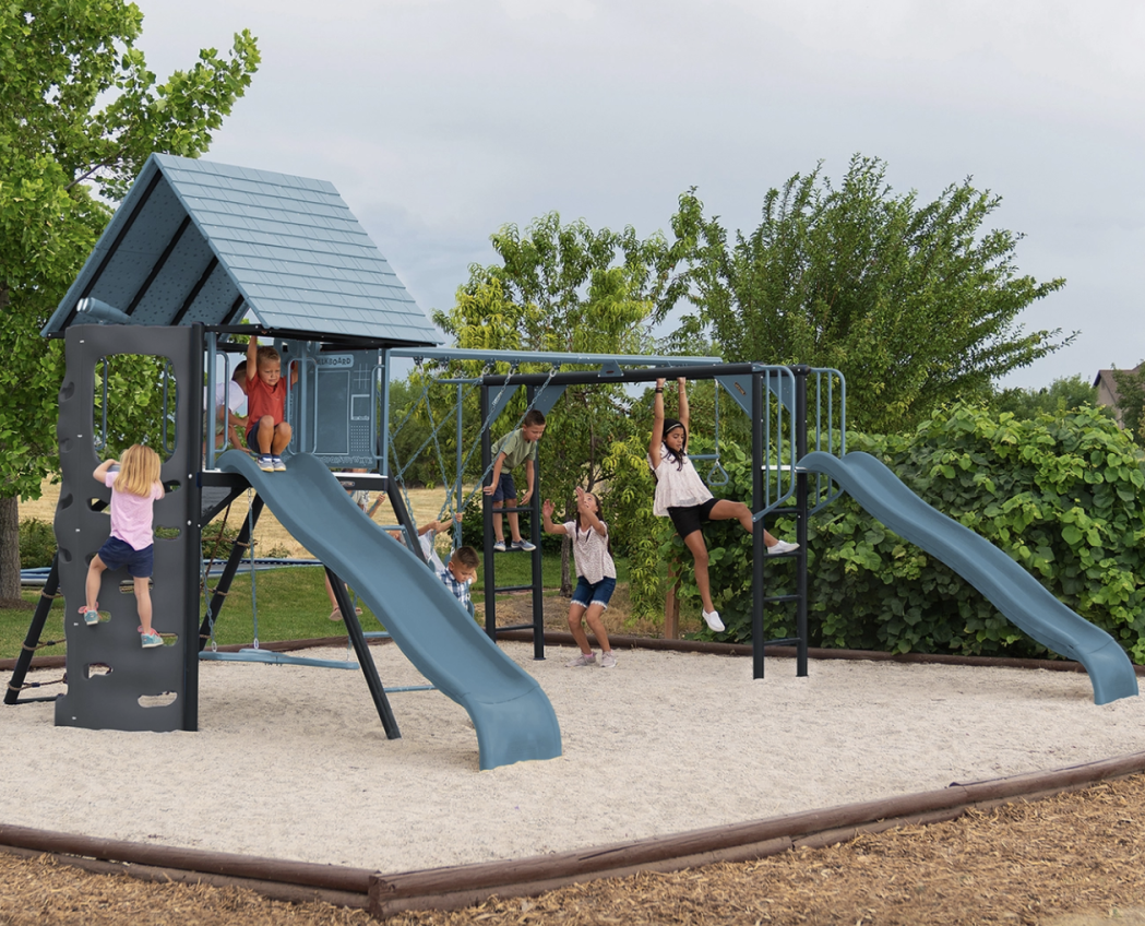 Children playing on a playground with slides, swings, and climbing structures on a sandy surface, surrounded by greenery and trees.
