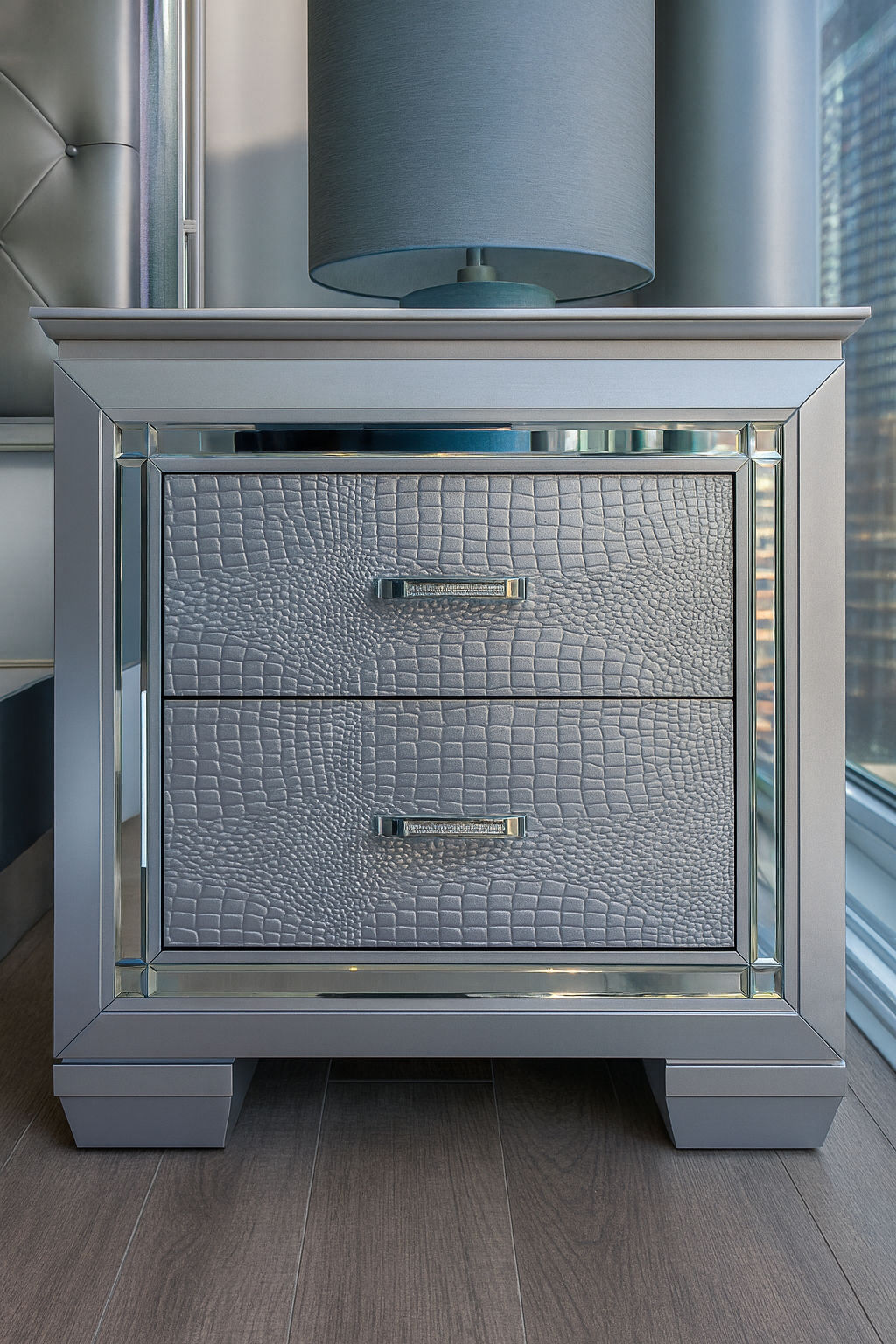 A silver nightstand with textured jewelry-like surface drawers and a glass top, with a gray table lamp on it, situated beside a window with blinds.