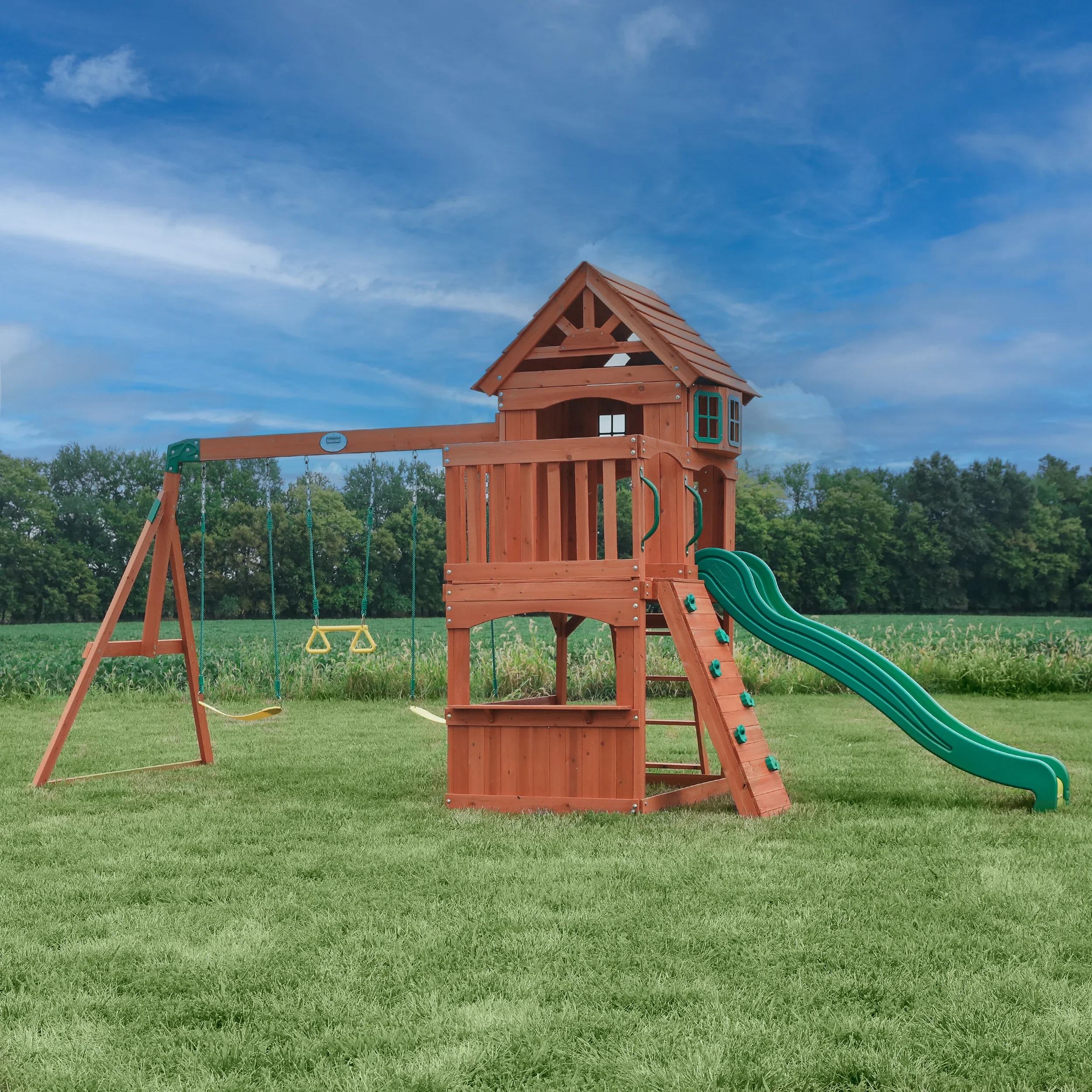 Wooden playset with slide, swings, climbing wall, and playhouse on a grassy field with trees and blue sky in the background.