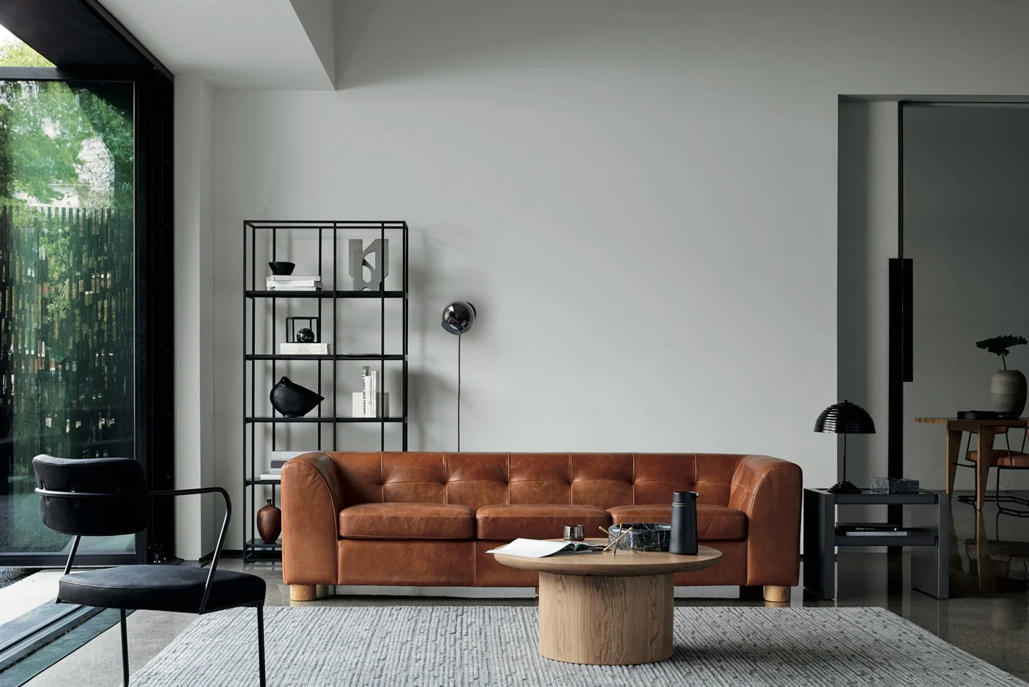 Modern living room with a brown leather sofa, black bookshelf, black armchair, round wooden coffee table, black side table with a lamp, and a large window revealing green outdoor scenery.
