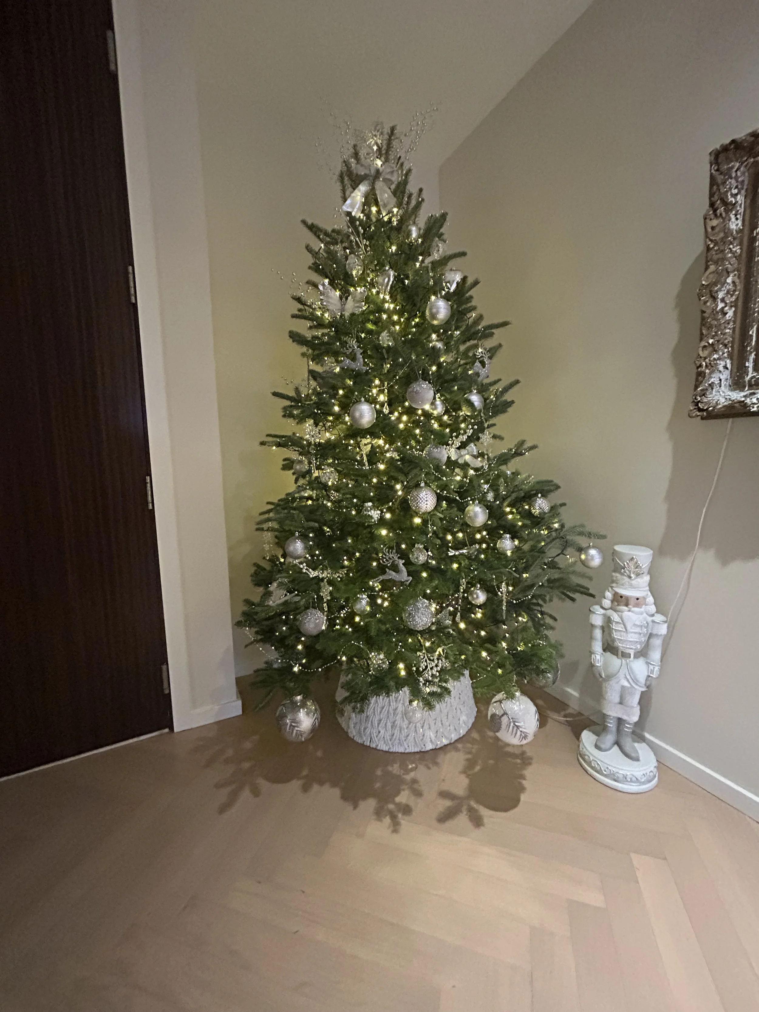 Decorated Christmas tree with white ornaments and lights, placed in a room corner next to a nutcracker figurine.