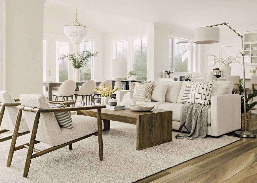 Bright, modern living room with white walls, large windows, a white sofa with pillows, a wooden coffee table, and armchairs, decorated with vases and plants.