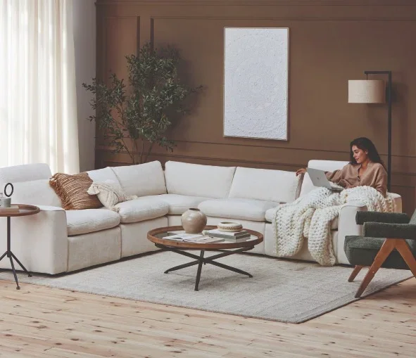Woman sitting on a white sectional sofa with a laptop in a modern living room, with a coffee table, tall tree, and wall art.
