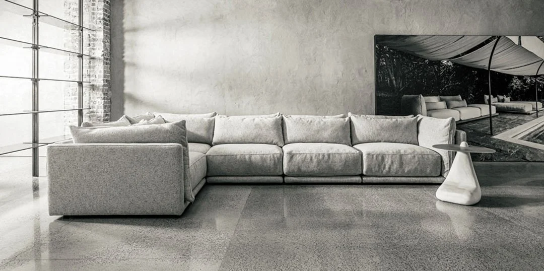 Light gray sectional sofa with matching cushions in a modern, industrial-style living room with a polished concrete floor and a wall with a large window.