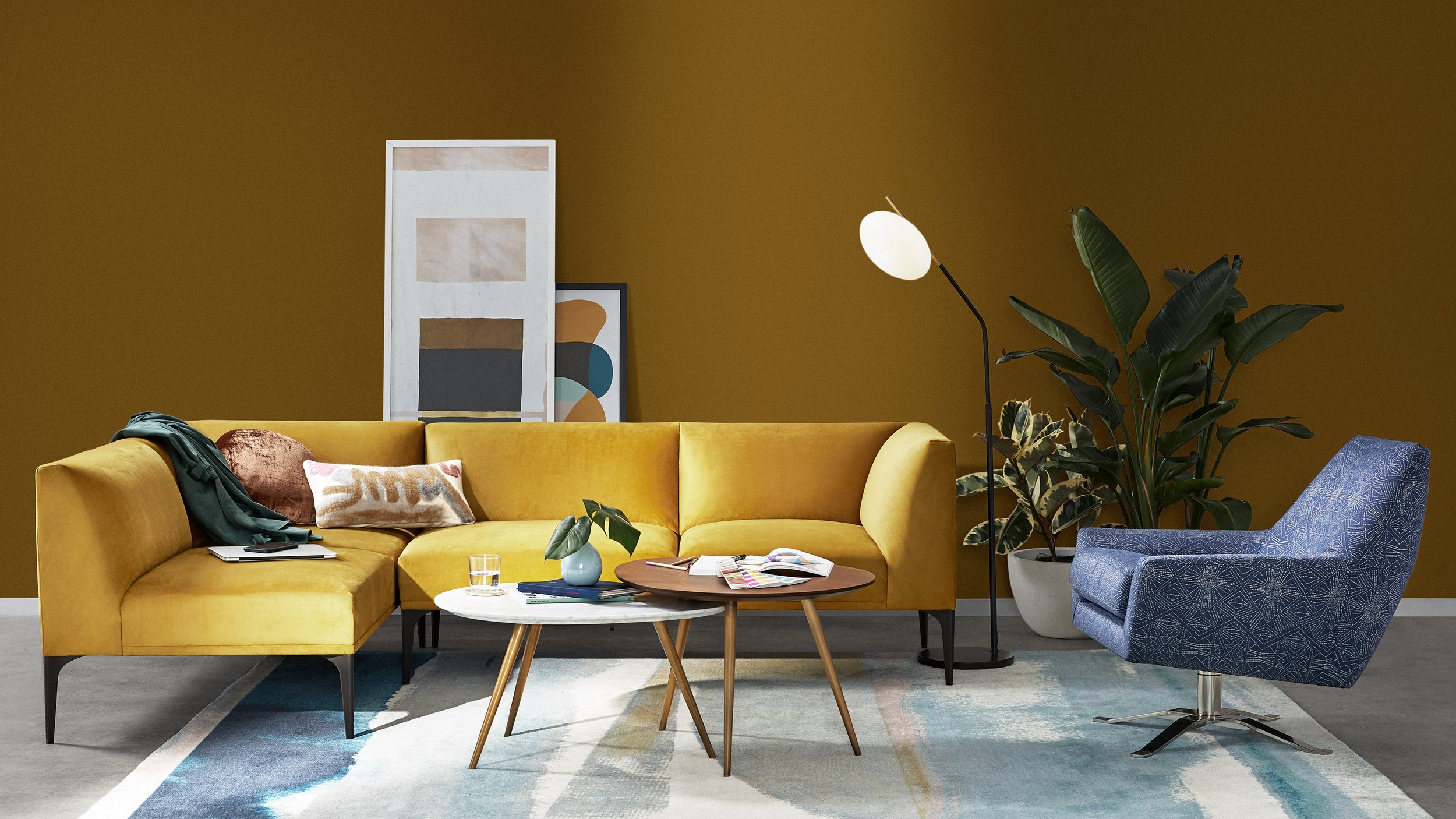 Living room with a mustard yellow sectional sofa, a blue patterned swivel chair, and two nested round coffee tables with books, a glass of water, and a small potted plant. Behind the sofa, two abstract art pieces and a tall indoor plant are visible. A floor lamp with a white shade is near the plant, and the wall is painted an earthy brown color.