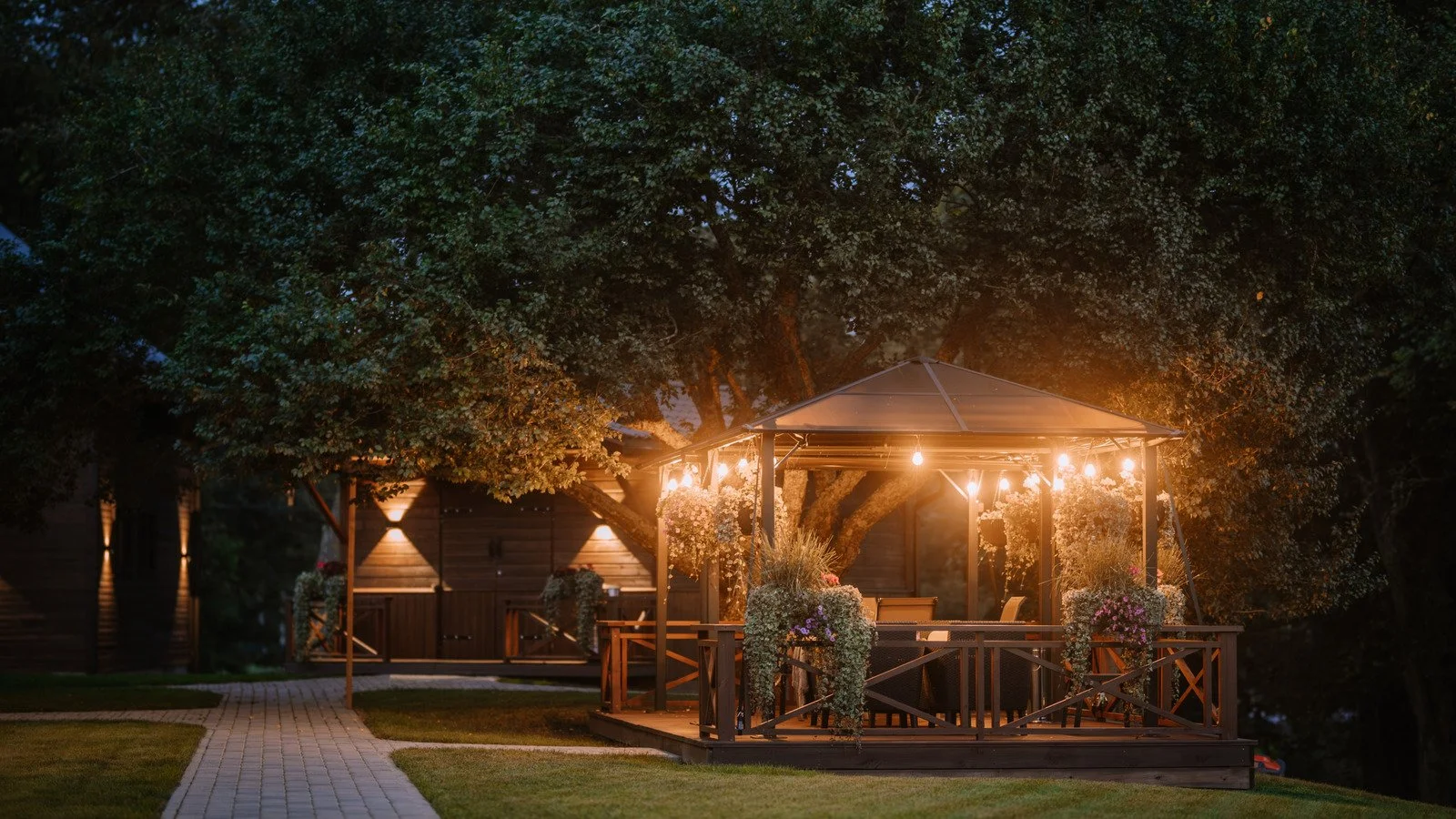 A lit outdoor gazebo surrounded by trees at dusk, with hanging string lights and potted plants on the railing, on a grassy lawn with a paved walkway.