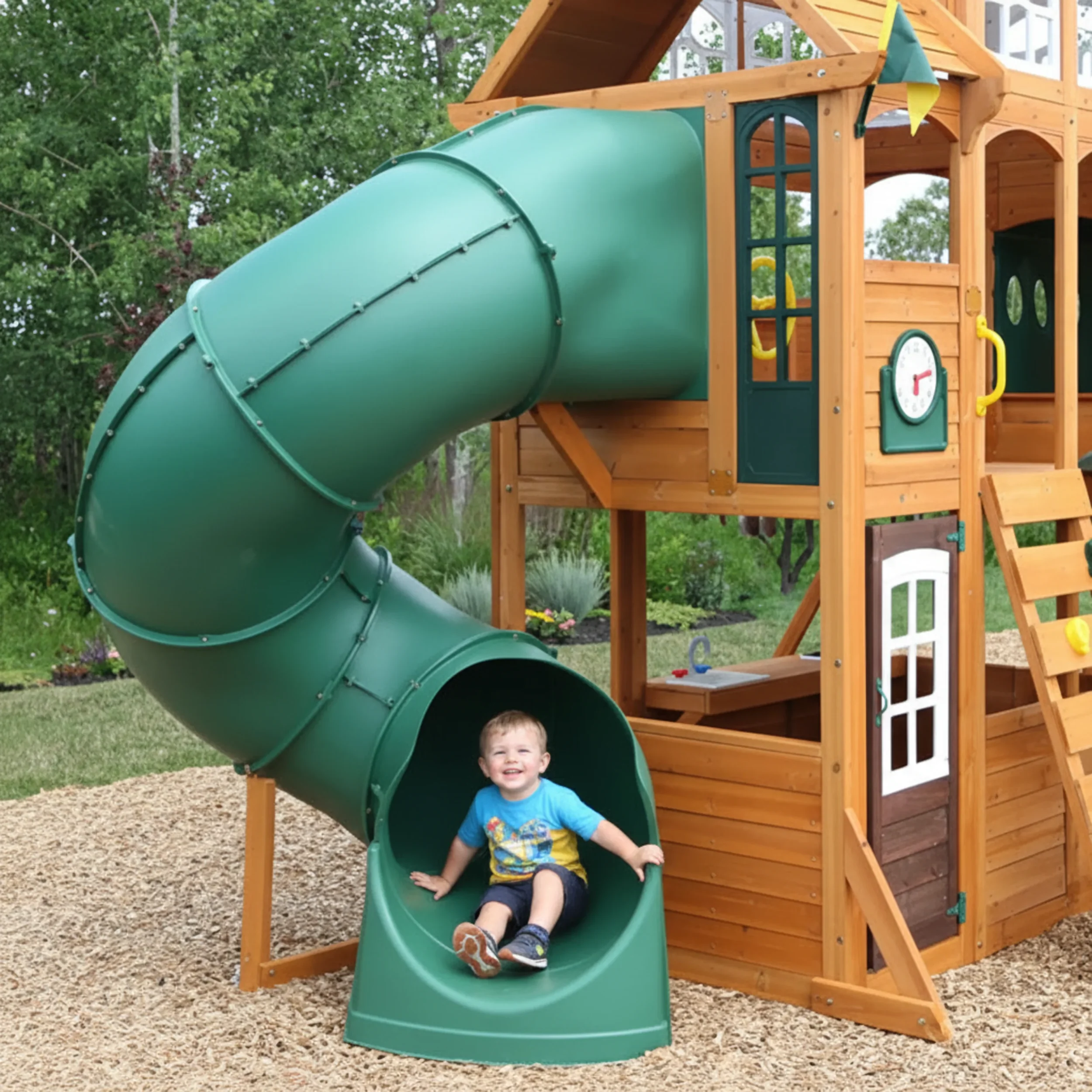 A young boy sitting at the bottom of a green tube slide attached to a wooden play structure in a backyard.