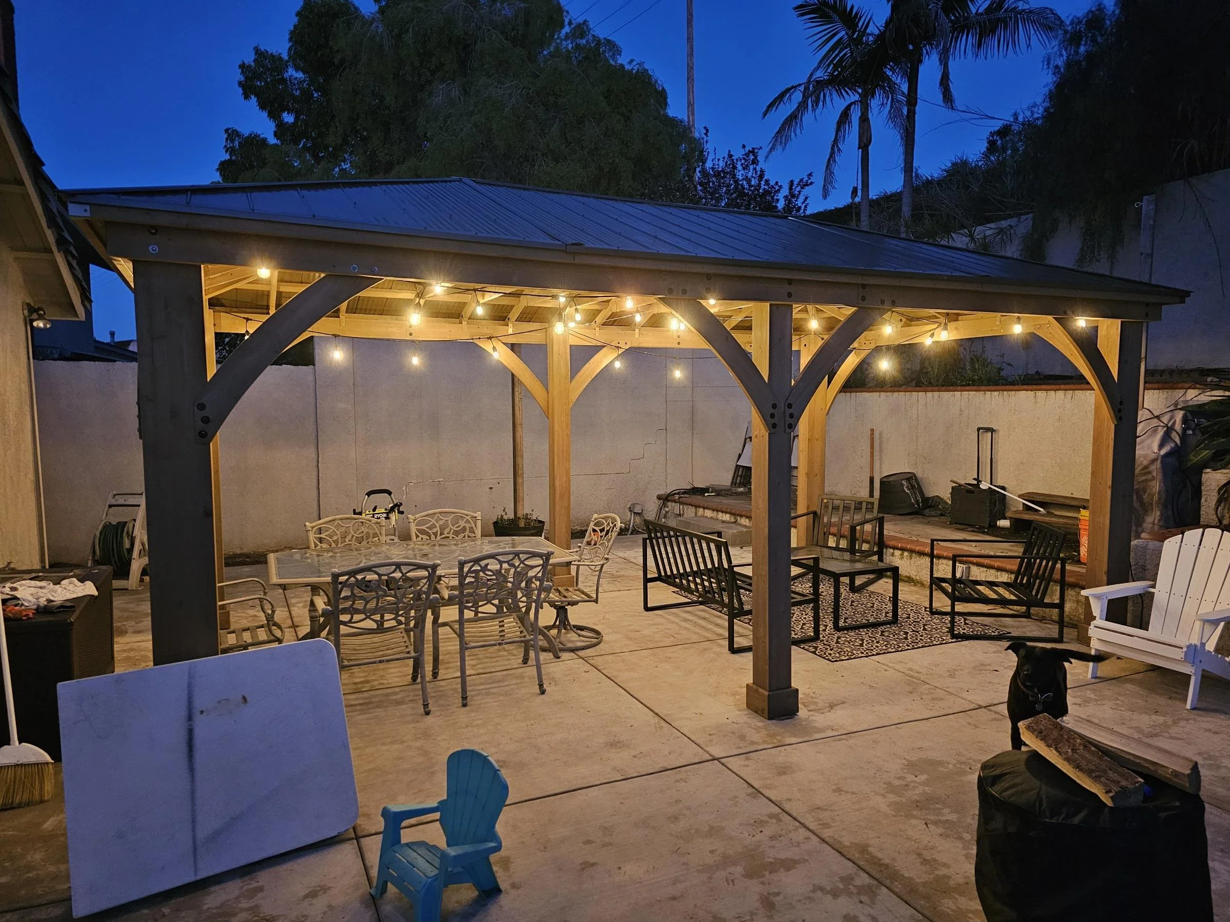 Nighttime backyard patio with a wooden gazebo decorated with string lights, patio furniture including chairs and tables, some stacked and some arranged, a black dog near a bag of firewood, and a white Adirondack chair, with trees and a blue sky in the background.