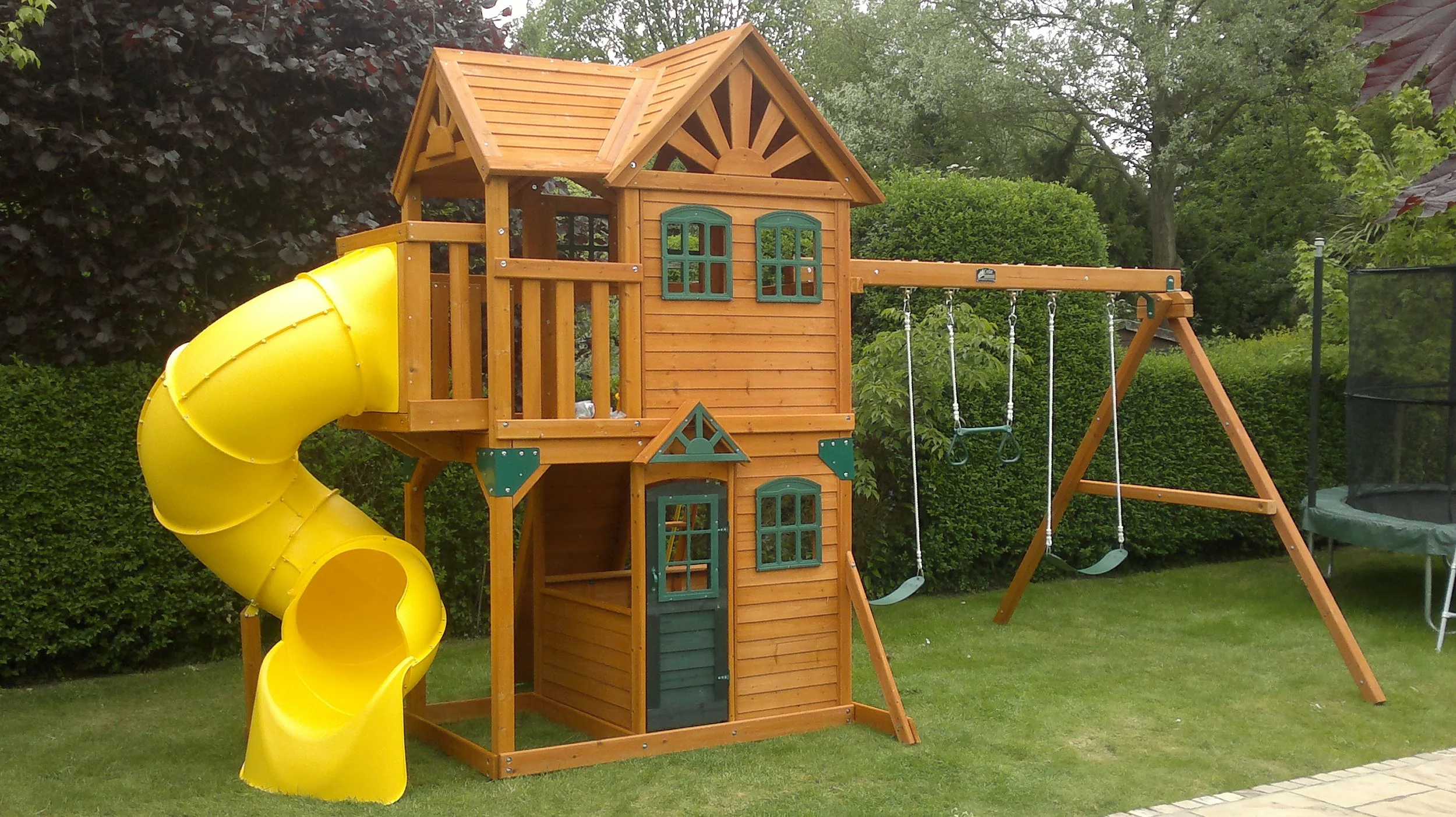 Wooden outdoor playhouse with a yellow spiral slide, swings, and a trampoline in a backyard with green grass and bushes.