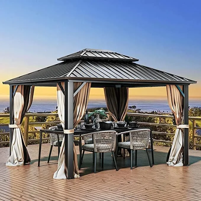 A gazebo with a metal roof and draped curtains, set on a wooden deck with a view of trees, a body of water, and a sunset in the background.