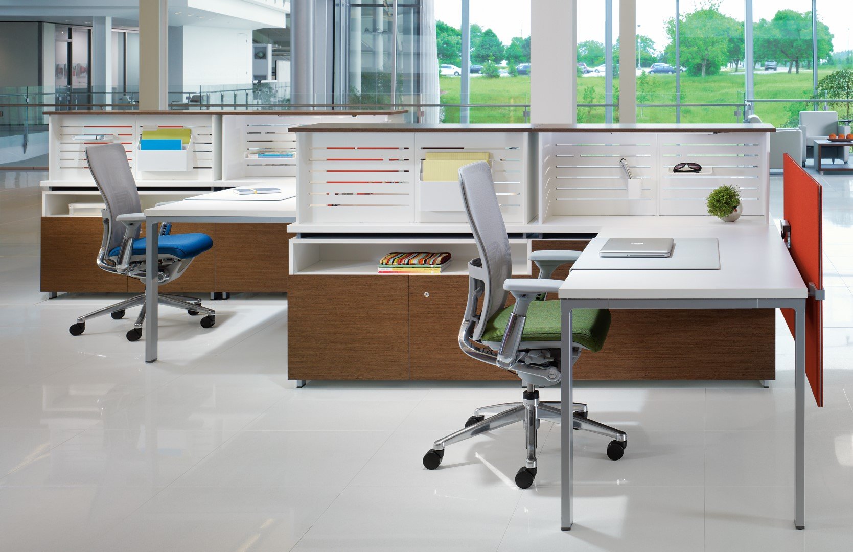 Modern office space with two white desks and ergonomic chairs, divided by a low white partition, with large floor-to-ceiling windows revealing green outdoor scenery.