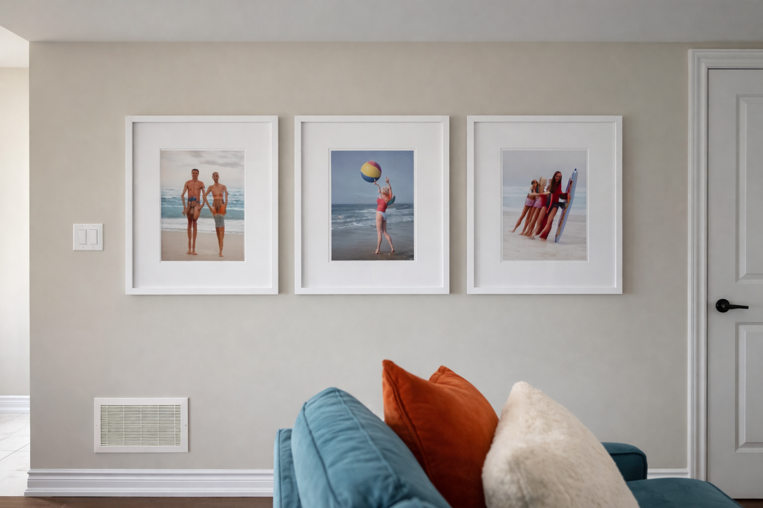 Three framed beach photos hanging on a beige wall above a blue couch with orange, white, and beige pillows.