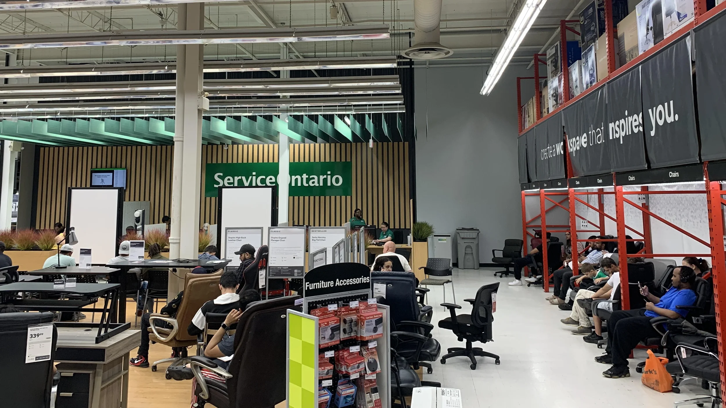 Furniture showroom with people sitting in chairs and waiting, featuring display sections for office chairs and furniture accessories, with a green sign reading 'Service Ontario' in the background.
