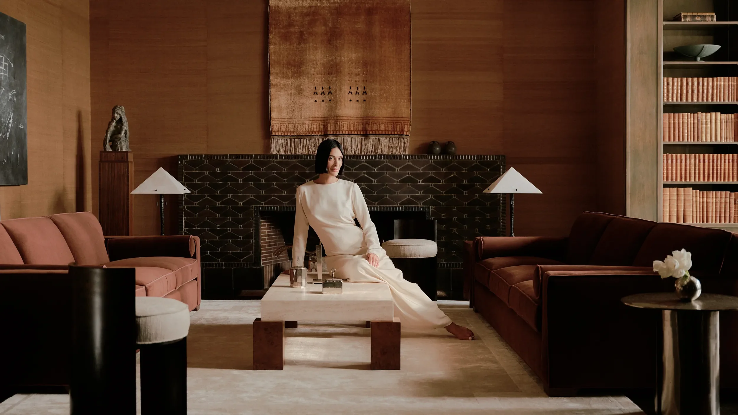 A woman in a cream-colored dress sitting on a bench in a sophisticated living room with wooden walls, a fireplace, a bookshelf, and various modern furniture and decor.