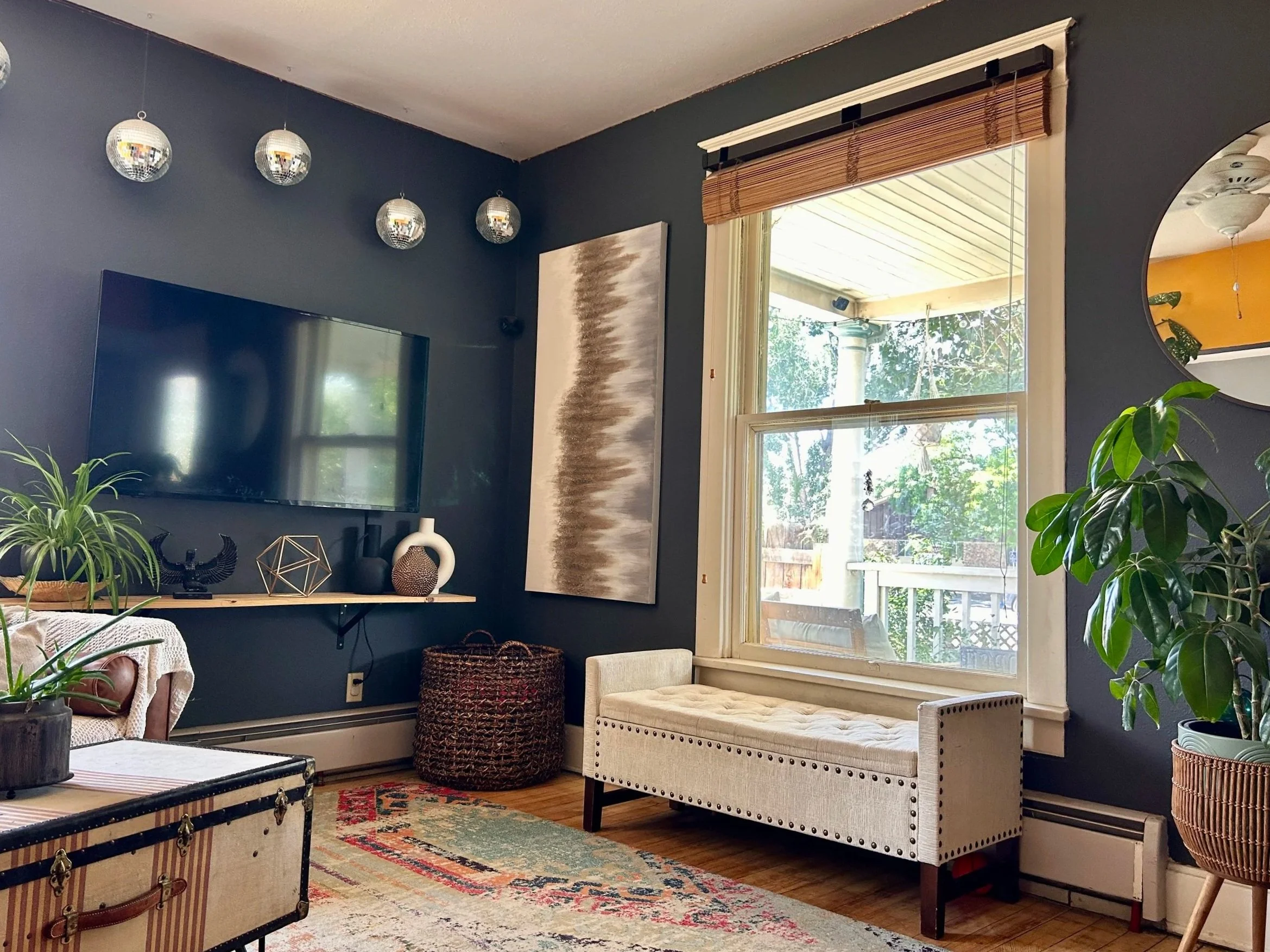 Living room with dark blue wall, hanging globe lights, flat-screen TV, abstract art, large window with bamboo shade, white upholstered bench, potted plants, vintage trunk, decorative objects, area rug, and ceiling fan.