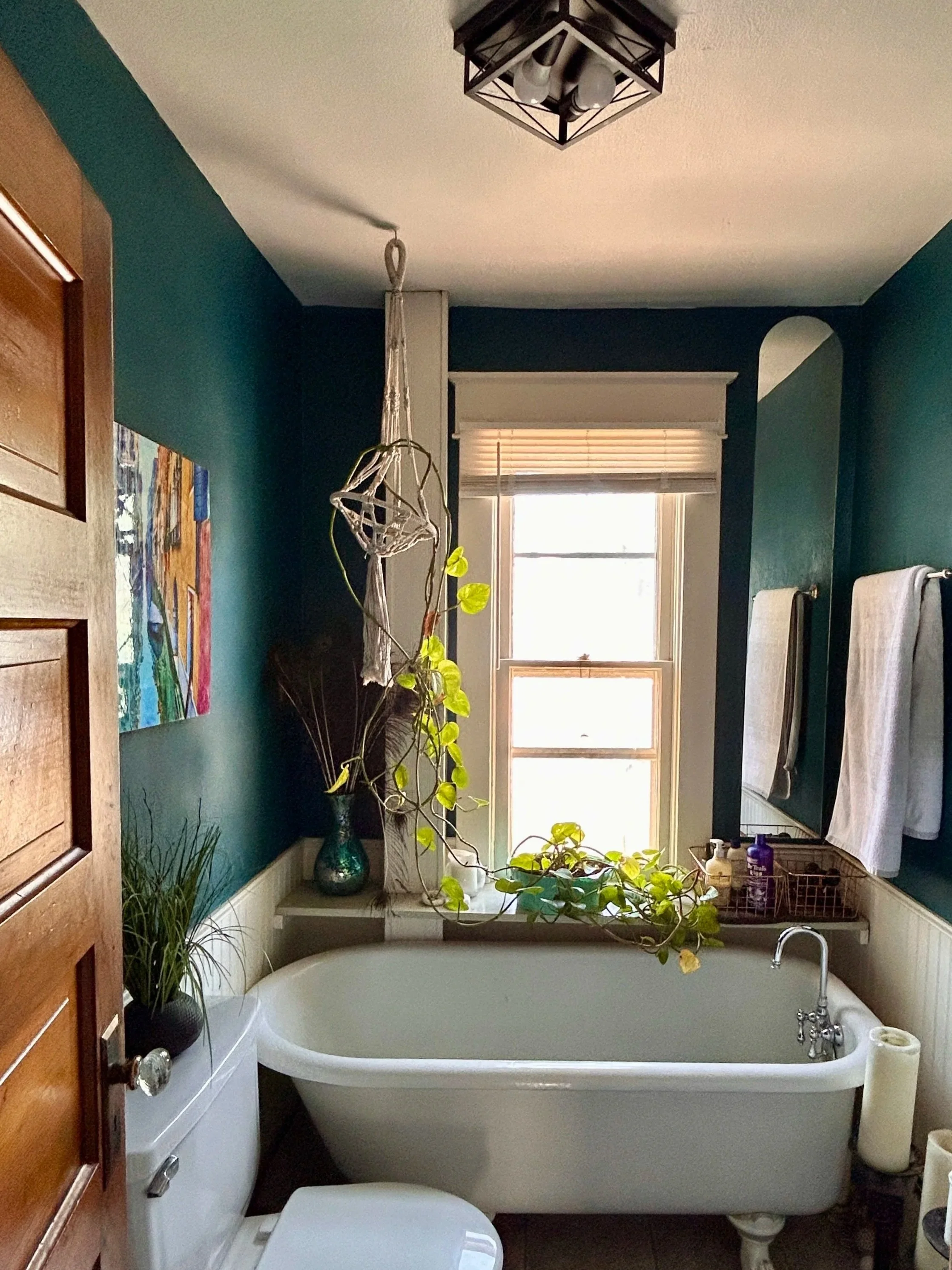 A bathroom with a bathtub, toilet, window, plants, and towels on a towel rack.