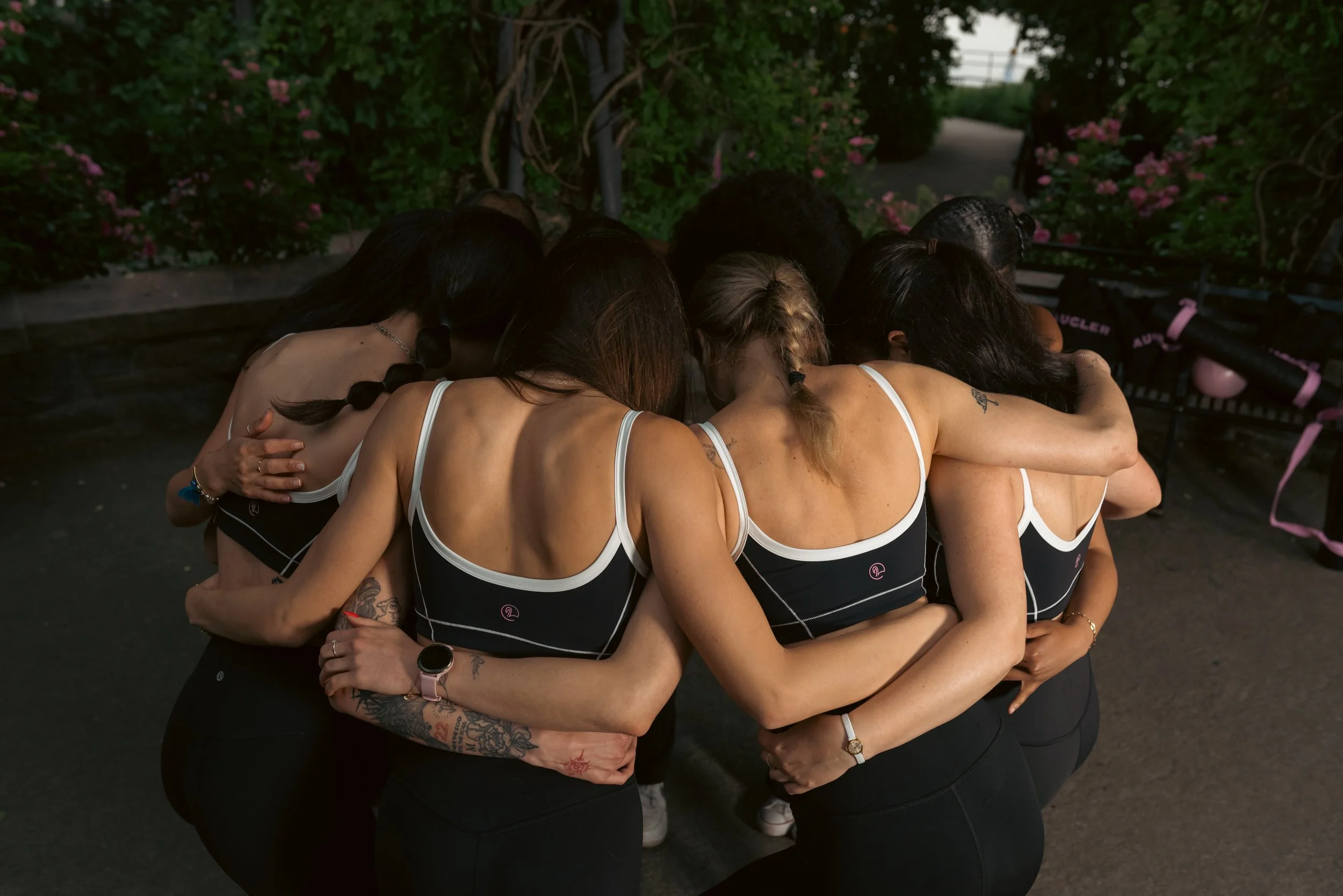 Group of women in a tight hug, wearing matching black and white athletic outfits, standing outdoors near greenery.