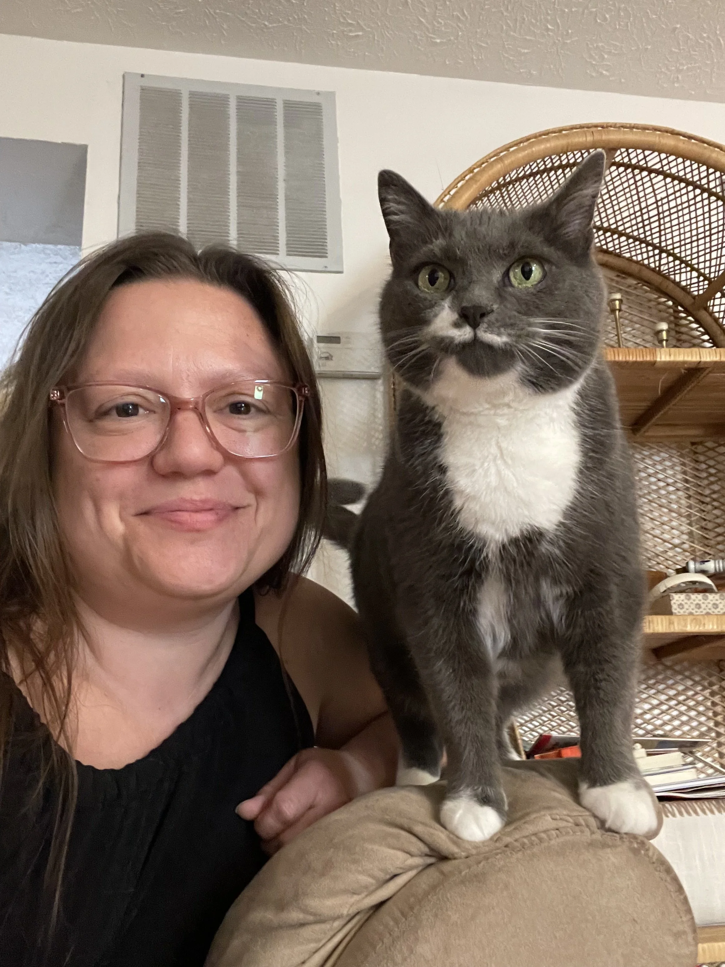 Woman with brown hair and glasses sitting happily next to a grey and white cat perched on a couch arm.