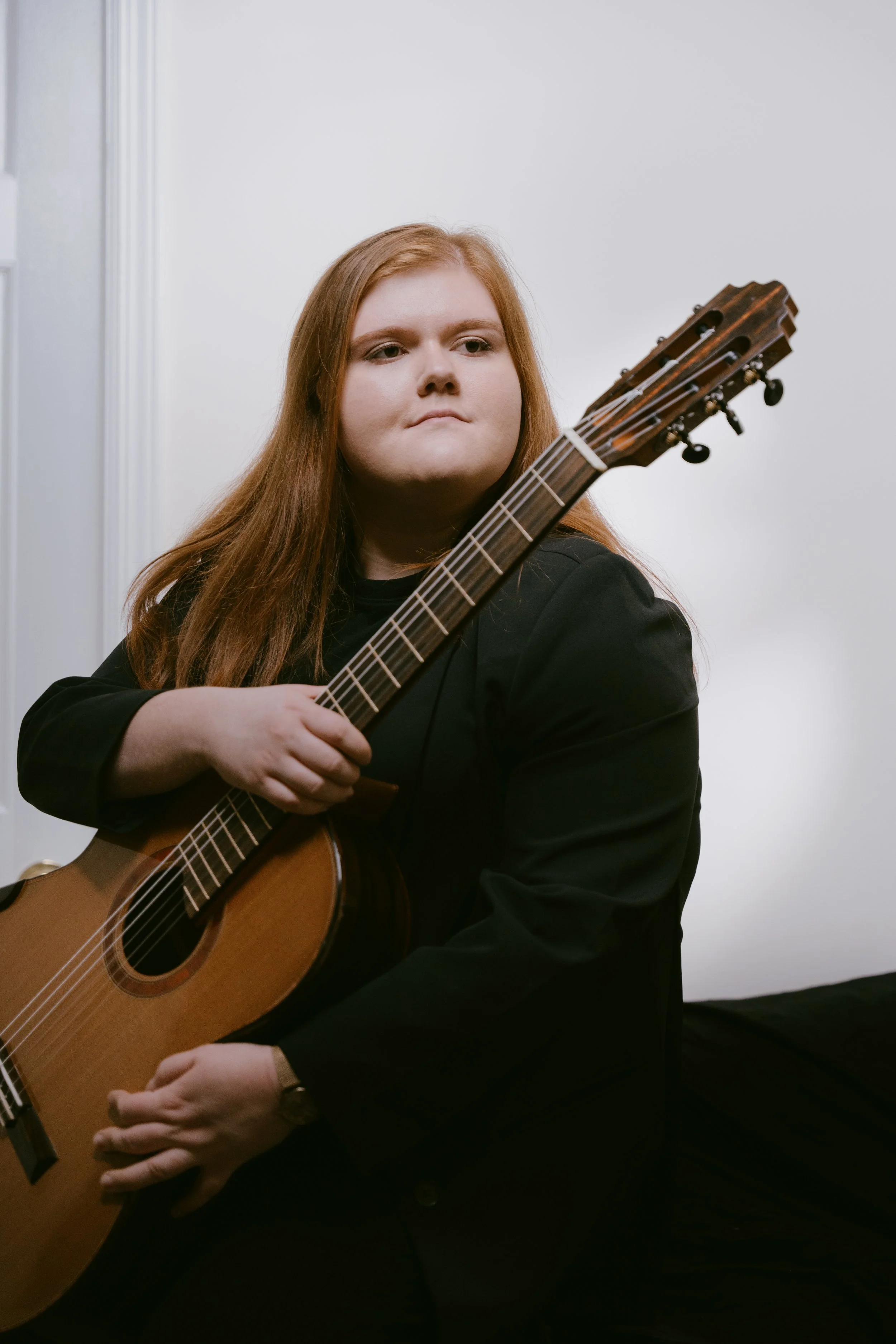 A woman with long red hair sitting with a guitar in her lap, wearing a black jacket, against a plain white wall.