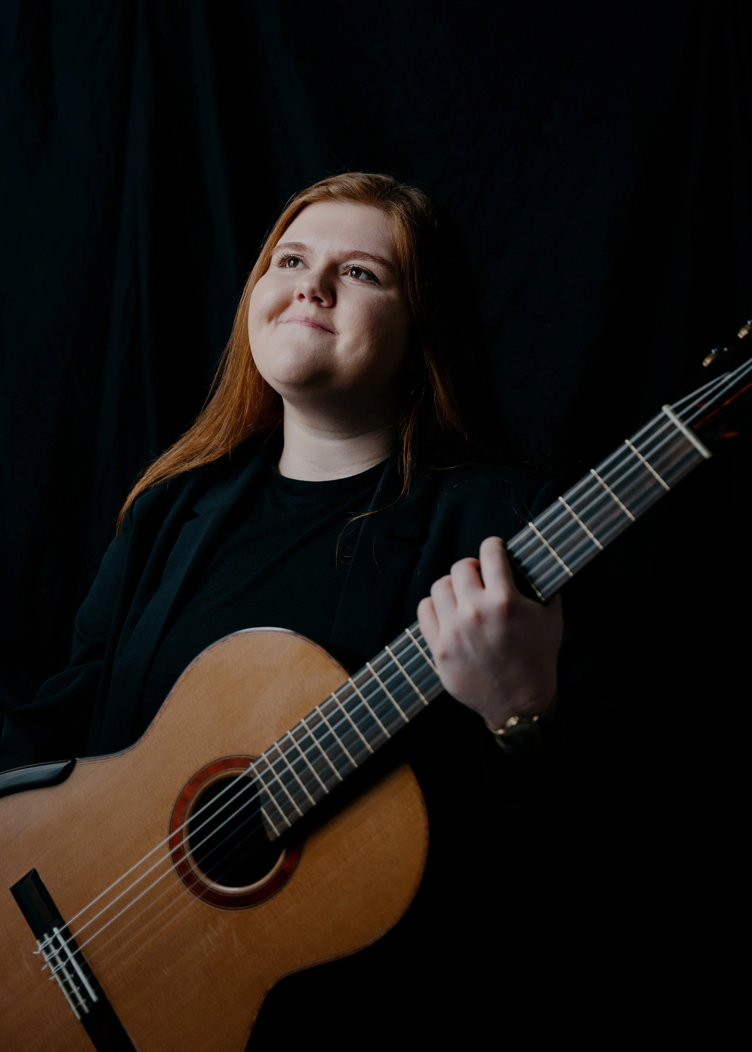 A woman with long red hair playing a classical guitar against a black background