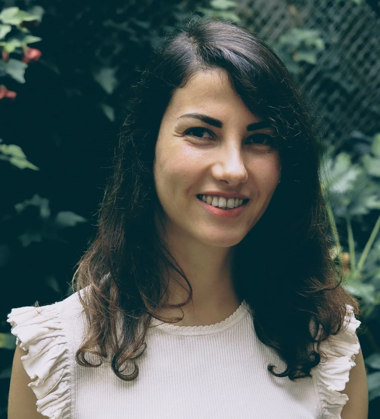 A woman with dark, wavy hair smiling outdoors with green foliage in the background.