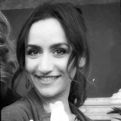 A woman with braided and beaded hair smiling, wearing earrings and a patterned top, in a setting with art or decorative items in the background.