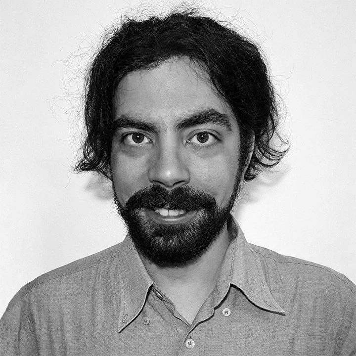 Black and white photo of a man with long curly hair, glasses, a beard, and a mustache, wearing a collared shirt and a necklace. He appears to be outdoors near a city with buildings and a mountain in the background.
