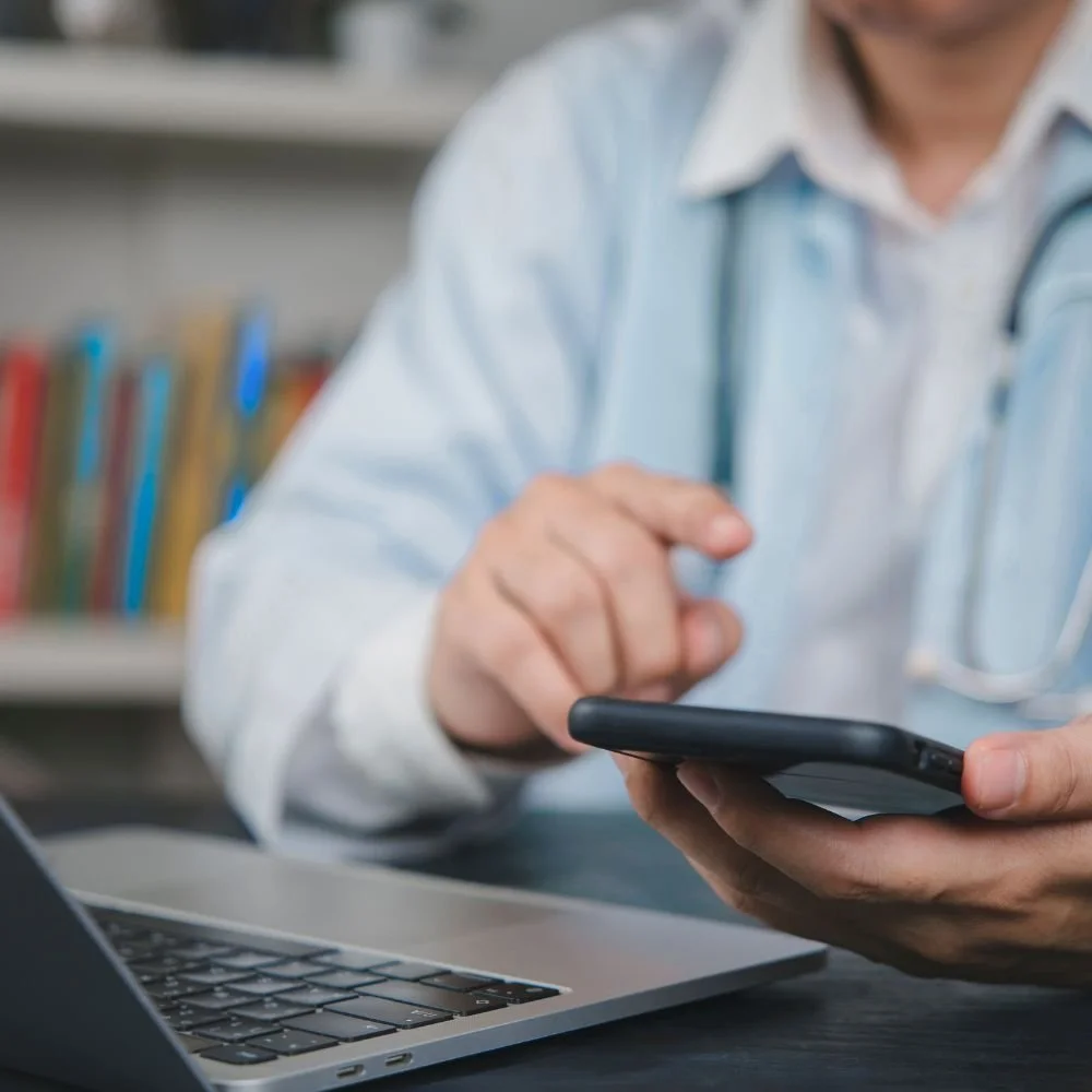 Health professional with stethescope around his neck sitting at a desk typing on his phone