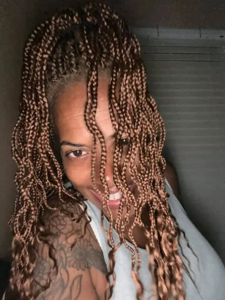 Close-up of a woman with tan-colored, twisted braids partially covering her face, smiling and showing her teeth, with a tattoo on her shoulder.
