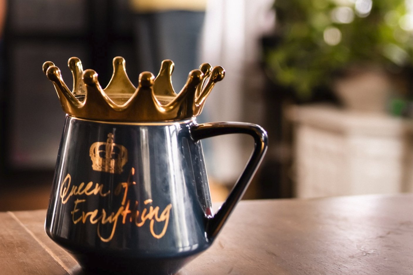 A black coffee mug with gold writing that says 'Queen of Everything,' topped with a small gold crown, placed on a wooden surface with a blurred background.
