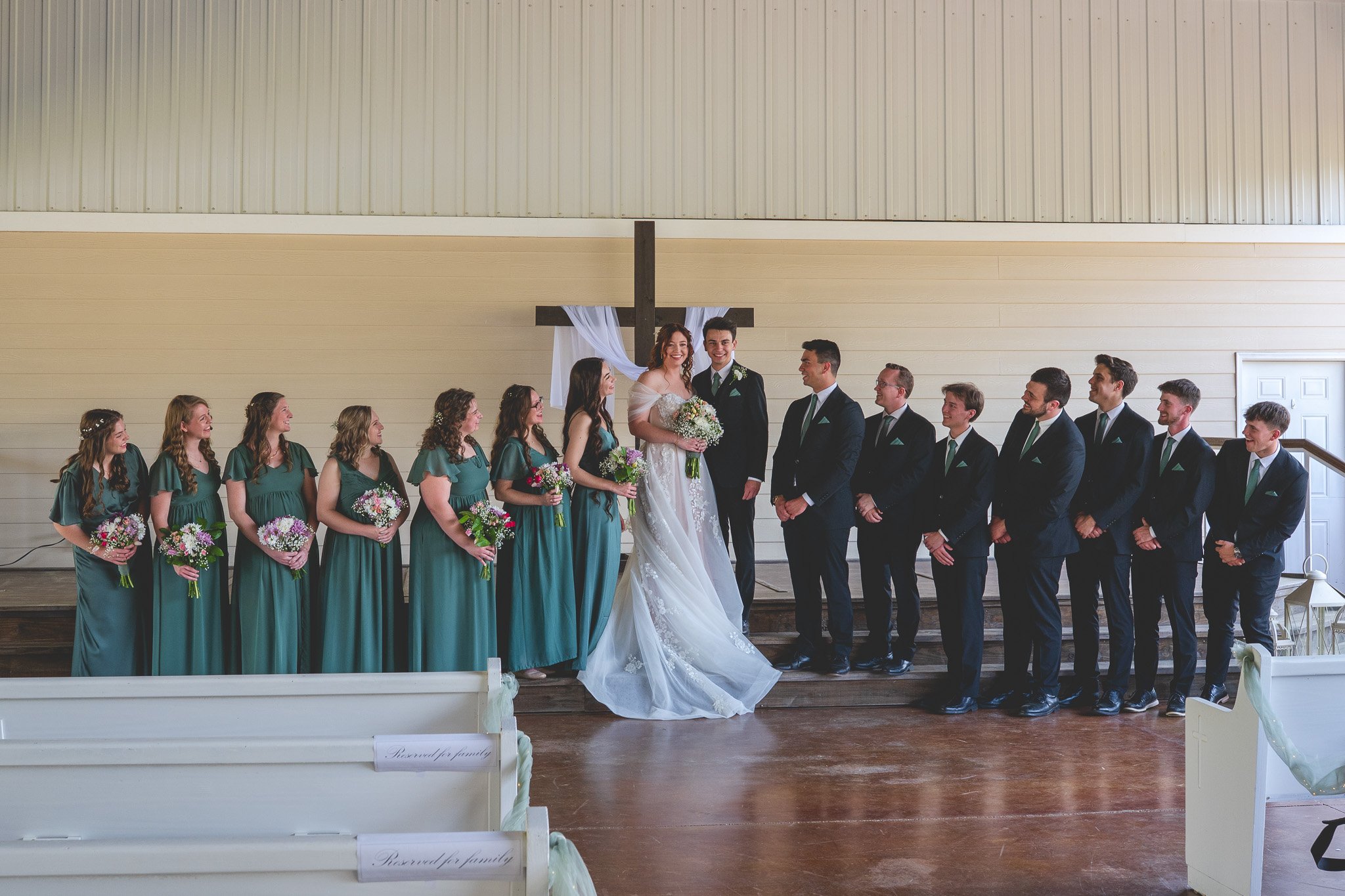 Wedding ceremony with bride and groom standing together in front of a cross, surrounded by bridesmaids and groomsmen, inside a church.